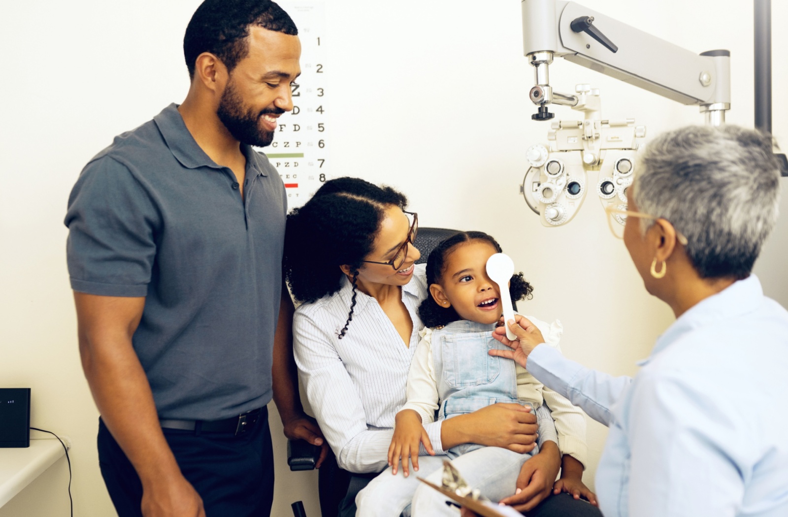 An optometrist covers a young girl's eye during an eye exam with the happy family nearby.