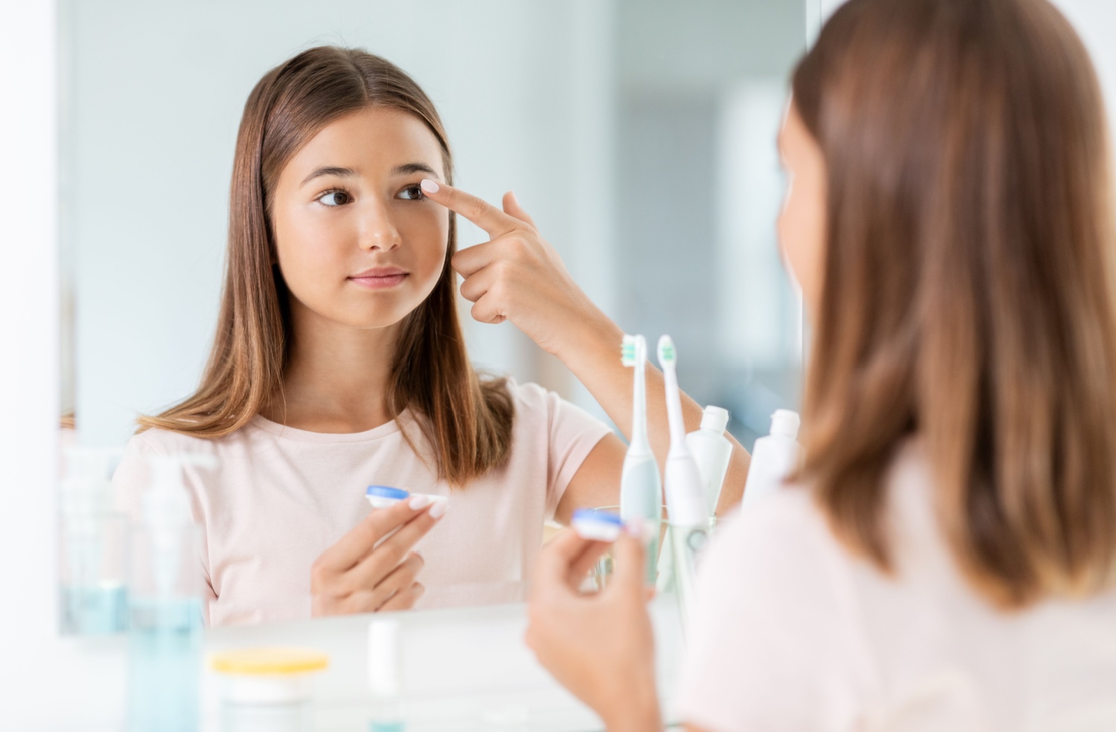 A teenager stands in their bathroom in front of a mirror. They are putting a prescription contact in their eye to help with their nearsightedness.