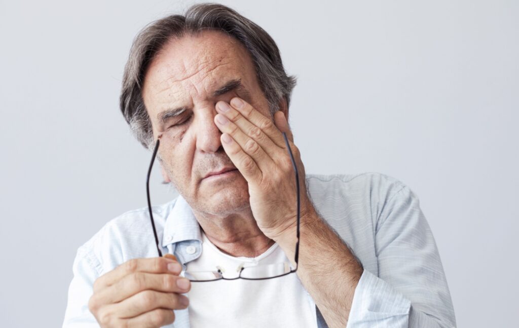 An older adult takes off their glasses and rubs their dry eyes to clear their blurry vision.