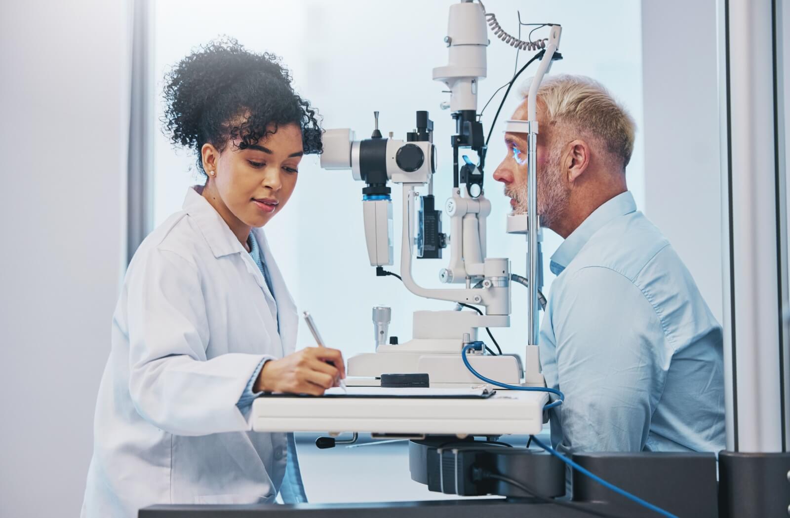 An optometrist in a white coat writing on a clipboard while assessing a patient's vision.